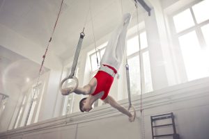 A male gymnast exercises on rings in an indoor gym, showcasing strength and flexibility.