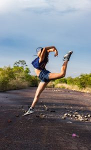 A woman performs a graceful dance leap on a sunlit outdoor path, showcasing fitness and flexibility.